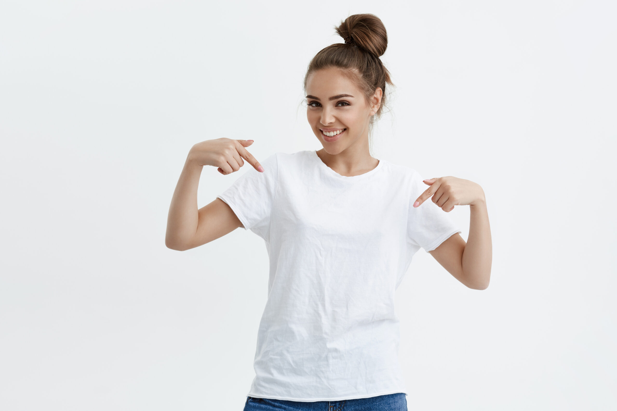 Charming emotive caucasian woman pointing down or at her t-shirt while smiling joyfully and expressing positive emotions over white background. Girl trained a lot, she wants to show her muscles.