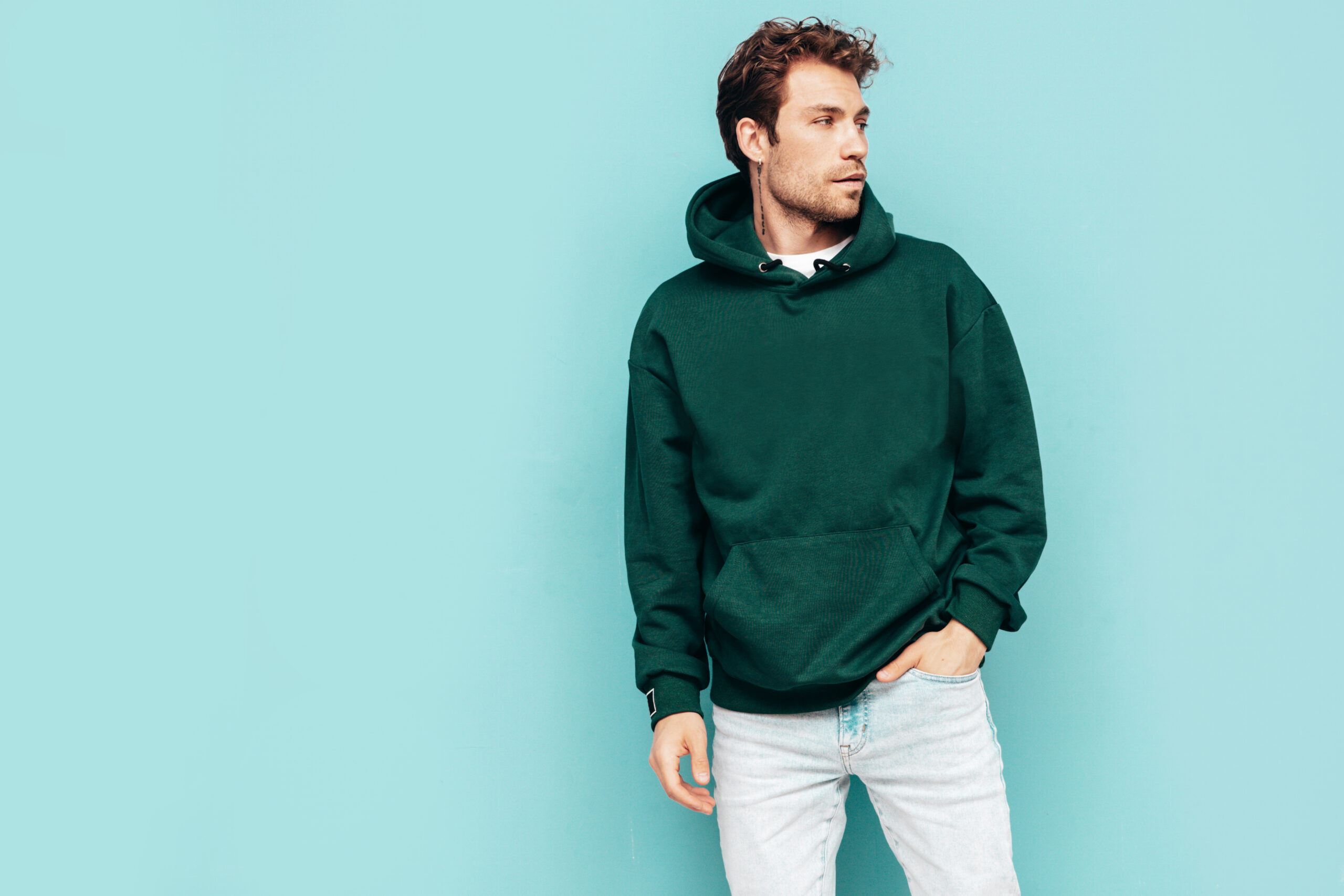 Handsome confident hipster model.Sexy unshaven man dressed in summer stylish green hoodie and jeans clothes. Fashion male with curly hairstyle posing in studio. Isolated on blue